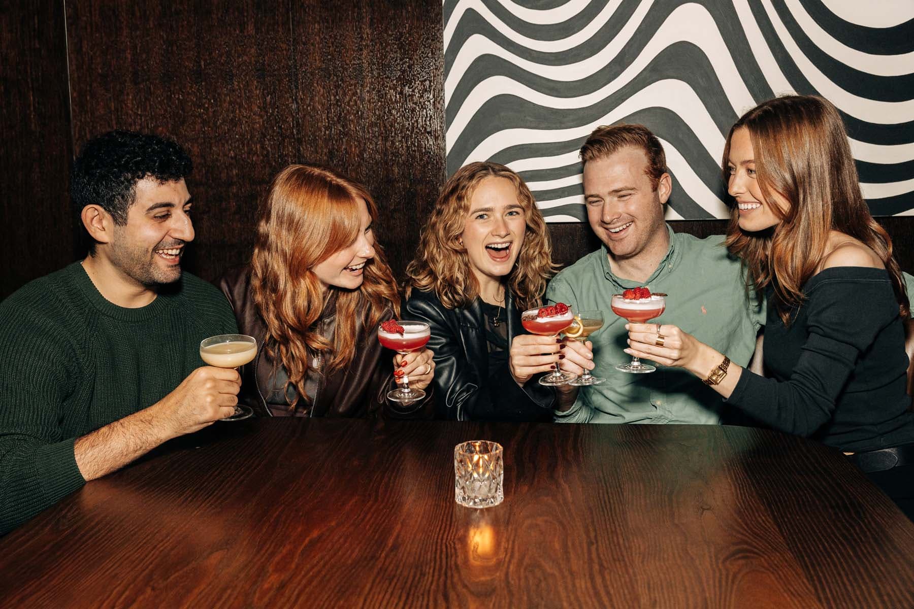 Five people at the end of a wooden table each holding a cocktail and raising their drinks in a toast.
