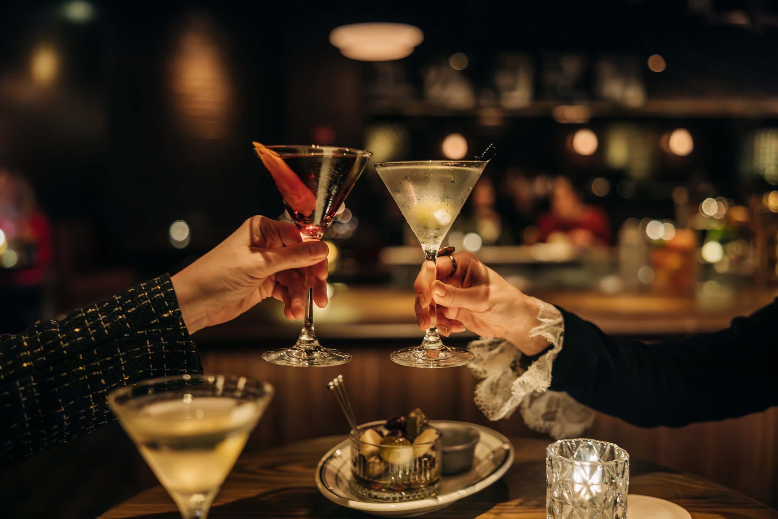 two hands, each holding cocktails served in martini glasses, coming together in a toast over a candle lit table.